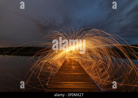 Ein dramatisches Langzeitbelichtungsfoto von sich drehender Stahlwolle, aufgenommen auf einem schmalen Holzsteg, der sich über einen ruhigen Landsee erstreckt. Stockfoto