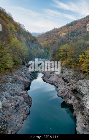 Ein schmaler Gebirgsfluss fließt durch eine dramatische Felsschlucht, die von steilen Hängen umgeben ist, die mit Herbstlaub bedeckt sind, das von Gelb und Orange bis d Stockfoto
