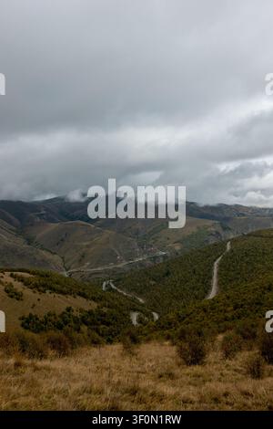 Eine weitläufige Luftperspektive zeigt ein tiefes Bergtal, das von einer schmalen Serpentinenstraße durchzogen ist, die sich durch bewaldete Hänge und offene Graslandschaften windet Stockfoto