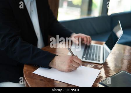 Kaukasischer Geschäftsmann schreibt Ideen auf Papier, während er in einem modernen Büro an einem Laptop arbeitet. Geschäftsplanung, Strategie und Produktivitätskonzept. Stockfoto