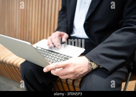 Nahaufnahme eines kaukasischen Geschäftsmannes, der im Freien auf einer Holzbank einen Laptop verwendet. Remote-Arbeit, mobiles Büro, Produktivität im Geschäftsleben und Technologiekonzept. Stockfoto