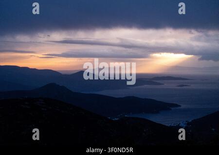 Schichtige Bergkämme, eine Meeresbucht und ferne Inseln erscheinen unter dunklen Gewitterwolken bei Sonnenuntergang in einer weiten Landschaftsansicht in Richtung Chora auf der Insel Amorgos Stockfoto
