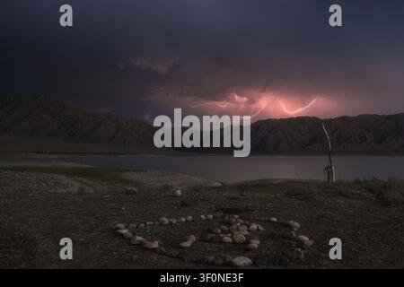 Entfernte Blitze, die bei Dämmerung über einem Bergsee einschlagen, mit einem Steinkreis am Ufer. Geeignet für Reise-Hintergründe, Abenteuerkonzepte Stockfoto