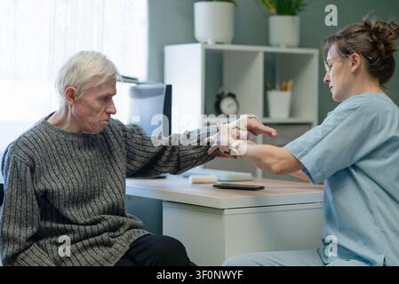 Krankenschwester unterstützt einen älteren Mann mit Knochenerkrankung und häufigen Knochenbrüchen im Krankenhaus. Gesundheitsfachkraft führt einen Senior mit verletztem Arm und Gips durch die medizinische Versorgung im Innenbereich Stockfoto