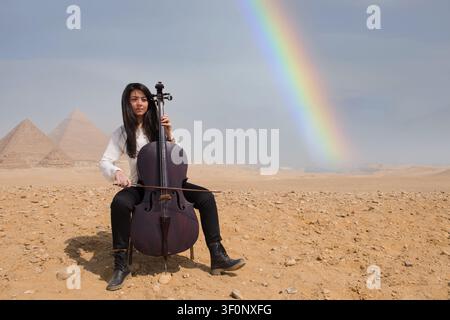 Junge arabische Cellistin spielt unter einem dramatischen natürlichen Regenbogen bei den ikonischen Pyramiden von Gizeh in der ägyptischen Wüste, ideal für Musik, Kunst und Kultur Stockfoto
