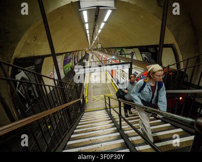 Passagiere steigen die Treppen von dem einzigartig schmalen Inselbahnsteig an der U-Bahn-Station Clapham North auf der Northern Line hinauf, während ein U-Bahn-Zug in South London, UK, wartet Stockfoto
