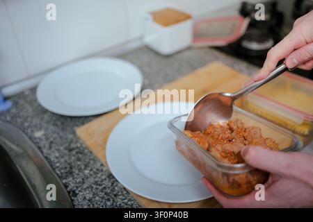 Hand schöpft gekochtes Schweinefleisch aus einem Glasbehälter, bereitet eine Mahlzeit auf einer Küchentheke zu Stockfoto