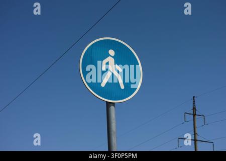 Ein rundes blaues Fußgängerweg-Verkehrsschild auf einem Metallpfosten vor einem blauen Himmel mit Stromleitungen. Stockfoto