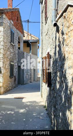 Eine schmale Stein-gasse, gesäumt von rustikalen Gebäuden mit Holzläden, Steinmauern und einem strahlend blauen Himmel darüber, der einen friedlichen Mittelmeerraum einfängt Stockfoto
