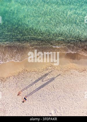 Luftaufnahme eines Paares, das sich an einem Sandstrand an den Händen hält, ihre langen Schatten erstrecken sich über den Sand neben türkisfarbenem Wasser während der goldenen Stunde in Sar Stockfoto