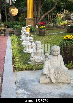 Reihe von steinernen kleinen Mönchsstatuen in einem friedlichen Tempelgarten. Bezaubernde Novizen-Buddha-Skulpturen säumen einen üppigen grünen Rasen. Bunte Blumen in Töpfen, t Stockfoto