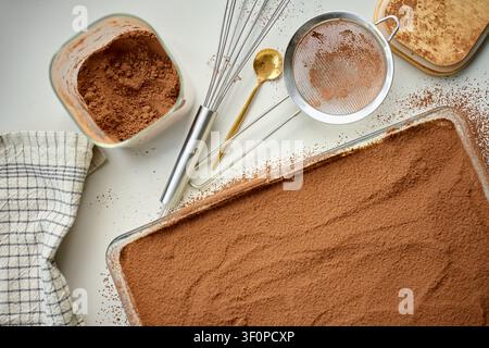 Kakaoverzogenes Tiramisu liegt in einer rechteckigen Glasschale neben einem Sieb und einer Kakaoschale auf dem Tisch. Konzept für hausgemachtes Dessert und die Zubereitung süßer Speisen Stockfoto