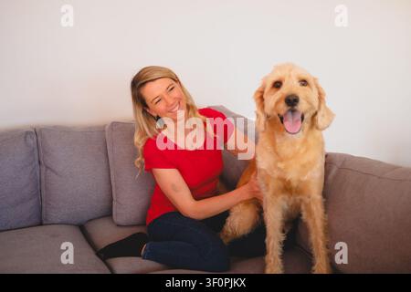 Frau lächelt, streichelt einen Goldendoodle-Welpen, der neben ihr auf einer grauen Couch sitzt Stockfoto