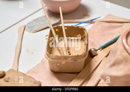 Nahaufnahme eines Behälters mit flüssigem Tonschlick in einer Töpferwerkstatt. Professioneller Handwerkerarbeitsplatz mit hölzernen Modellierwerkzeugen und rohem Ton auf weißem Hintergrund Stockfoto