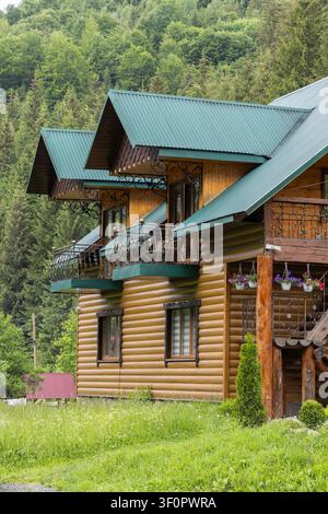 Ein mehrstöckiges Holzblockhaus mit Eisenbalkonen steht an einem steilen Hang, der mit dichtem Nadel- und Laubwald bedeckt ist. Stockfoto