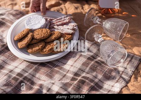 Ein weißer Teller hält Schokoladenkekse und Käse-Sticks auf einer karierten Decke. Zwei Kristallweingläser und eine Weinflasche stehen in der Nähe an einem Sandstrand Stockfoto