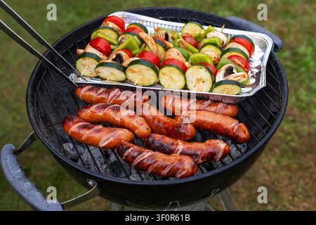 Gegrillte Würste und Gemüsespieße auf dem Grill. Gegrillte Würste garen auf einem Holzkohlegrill neben bunten Gemüsespießen mit Zucchini Stockfoto