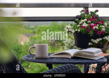 Augenhöhe-Bild, das eine weiße Tasse und ein aufgeschlagenes Buch auf einem schwarzen Metalltisch zeigt, mit hängendem Blumenkorb und verschwommenem grünen Baumhintergrund Stockfoto