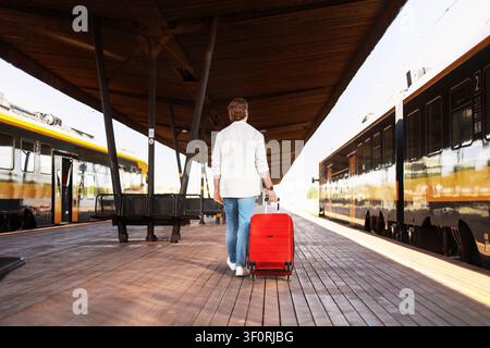 Ein Passagier geht auf einem Bahnhofsgleis. Der Mann zieht einen roten Koffer und rollt sein Gepäck für eine sonnige Reise. Hochwertiges Foto Stockfoto