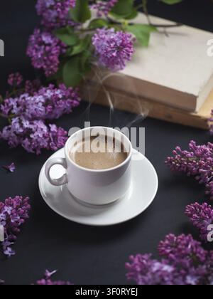 Eine weiße Tasse duftenden Kaffees auf einem dunklen Tisch mit Fliederstrauch und einem Buch. romantische Wohnatmosphäre Stockfoto