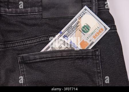 Paar schwarze Jeans mit einem Hundert-Dollar-Schein, der aus der Gesäßtasche schaut, betont die abgenutzte Denimtextur und die klaren Geldscheindetails, minimalistisches Set Stockfoto