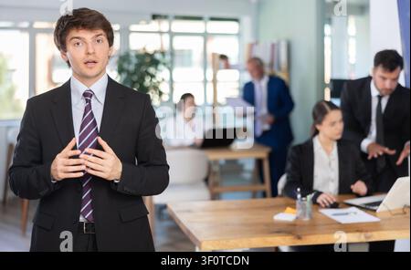 Positiver junger Geschäftsmann in Business-Kleidung vor dem Hintergrund eines Kollegen, der in einem Firmenbüro arbeitet Stockfoto