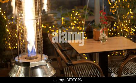 Leere Restaurantterrasse bei Nacht mit leuchtendem Gas-Brennofen, Bistrostühlen und Holztablett hinter Windschutz, warmes Feenlicht-Bokeh, Außengarten Stockfoto