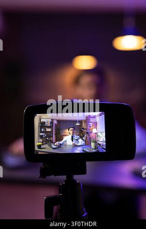 Asiatischer Mann führt Online-Podcast in seinem Heimstudio, spricht in ein Mikrofon mit Laptop und Audiogerät. Nahaufnahme zeigt Kamera, die den männlichen Vlogger bei der Erstellung von Inhalten aufnimmt, der Geschichten teilt und das Publikum einbindet. Stockfoto