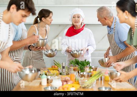 Erwachsene Schüler bereiten Speisen nach Rezept in einem Kochkurs zu, der von einer Küchenchefin geleitet wird Stockfoto