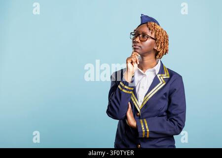 Nachdenkliche Flugbegleiterin in Uniform steht mit der Hand am Kinn und blickt nachdenklich vor einem blauen Hintergrund. Flugbegleiterin plant und trifft Entscheidungen für ihre Laufbahn in der Luftfahrt mit konzentriertem Ausdruck. Stockfoto