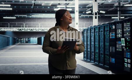 Der Systemtechniker mit einem digitalen Tablet führt durch die Serverreihen der Servereinrichtung und führt fortschrittliche Energiemanagementlösungen ein. Stockfoto