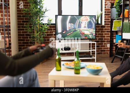 Gruppe von Freunden spielt zu Hause Videospiele, hält Controller und lehnt sich zum Fernseher. Junge Leute sitzen mit Getränken und Snacks, genießen Freizeit und Unterhaltung in einem gemütlichen Wohnzimmer. Stockfoto