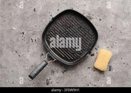 Schmutzige Gusseisen-Grillpfanne mit verbrannten Lebensmittelrückständen und einem Reinigungsschwamm auf grauem Betonhintergrund, daneben liegt ein gelber Reinigungsschwamm Stockfoto