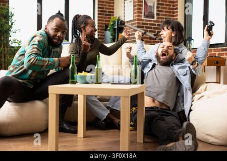 Multikulturelle Gruppe von Freunden feiert den Sieg beim Videospiel in einer gemütlichen Wohnung. Fröhliche, verschiedene Personen verbinden sich über Getränke, Spiele und Lachen, während sie Freundschaft und Wochenend-Auszeit genießen. Stockfoto