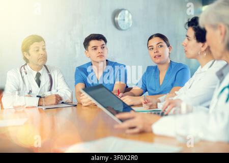 Ärzte und Krankenschwestern unterschiedlichen Alters diskutieren das Tagesthema, während sie mit Laptop und Notizen am Tisch sitzen Stockfoto