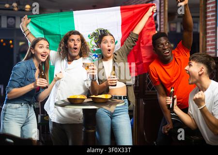 Fröhliche multirassische Sportfans, die mit der Flagge Mexikos wedeln, während sie in der Sportbar Bier trinken Stockfoto