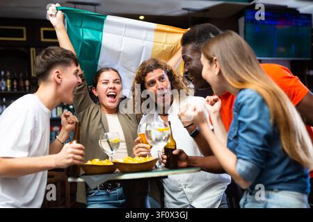 Fröhliche, multirassische Fußballfans schwenken die Flagge Irlands, während sie in der Sportbar Bier trinken Stockfoto