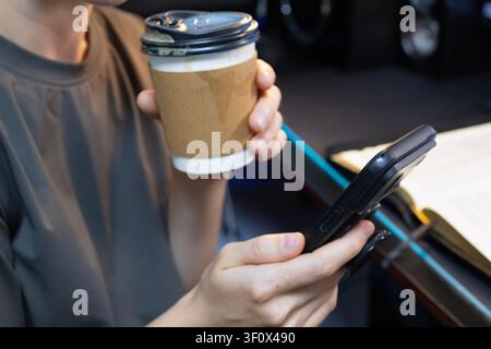 Junge Frau benutzt Smartphone und hält eine Kaffeetasse, während sie remote arbeitet Stockfoto