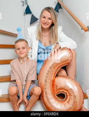 Mutter und Sohn sitzen auf einer Treppe mit Ballons, in einer Innentreppe, mit Ballons, Treppe, mit Textil, hölzerner Geländerstruktur, von vorne Stockfoto