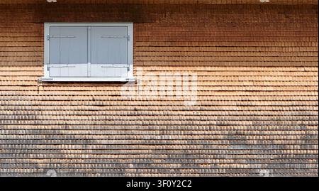 Verwitterte Holzdachplattenhauswand mit geschlossenem Fenster und weißen Fensterläden im ländlichen Schweiz Stockfoto
