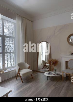 Hell minimalistisches Wohnzimmer mit einem großen Spiegel, einem gemütlichen Stuhl und einem schneebedeckten Fensterblick. Stockfoto