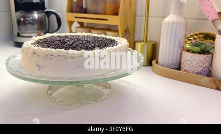 Hausgemachter Schokoladenkuchen mit weißer Glasur und Schokoladenspritzeln auf einem Glasständer in einer Küche Stockfoto