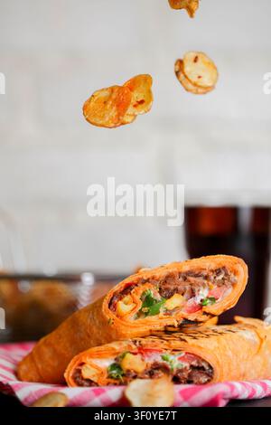 Zwei Wrap mit Fleisch und Gemüse stehen auf einem Tisch. Kartoffelchips fallen von oben. Ein Getränk steht in der Nähe. Die Szene zeigt die Zubereitung von Speisen und Genuss Stockfoto