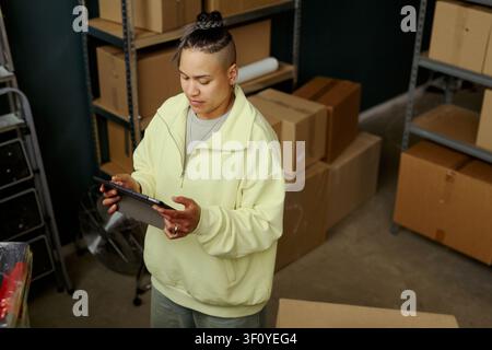 Hochwinkelaufnahme einer jungen erwachsenen biracialen Frau, die ein Tablet überprüft, während sie das Paketlager in einem Lagerhaus verwaltet, um die Auftragserfüllung und das Marketing für kleine Unternehmen zu unterstützen Stockfoto