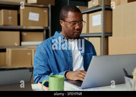 Mittlere Aufnahme eines afroamerikanischen jungen Erwachsenen, der in einem Lagerbüro die Inventarverwaltung auf einem Laptop durchführt, um E-Commerce-Operationen und Logistik-Marketing für kleine Unternehmen zu unterstützen Stockfoto