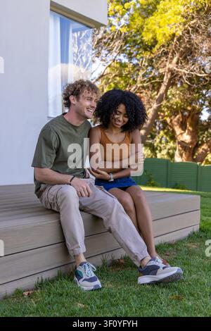 Verschiedene Paare sitzen auf einer erhöhten Holzterrasse im Hinterhof, tragen Turnschuhe und Jeansrock. Paar, Erwachsene, Terrasse, Terrasse, Rasen, Hof, sonnendurchflutete Stockfoto