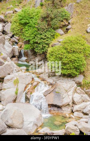 Ein kleiner Wasserfall stürzt über große graue Felsen, umgeben von üppigen grünen Büschen in den Himalaya-Bergen. Stockfoto