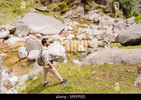 Ein männlicher Wanderer mit einem Rucksack steht an einem grasbewachsenen Ufer mit Blick auf einen felsigen Gebirgsbach im Himalaya. Stockfoto