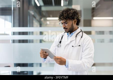 Junger männlicher Arzt mit Stethoskop, der einen weißen Laborkittel trägt, konzentriert sich auf ein digitales Tablet in einer modernen Klinik oder im Krankenhaus, repräsentierend Gesundheit Stockfoto