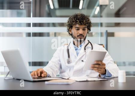 Junger männlicher Arzt mit lockigen Haaren, der einen weißen Laborkittel und ein Stethoskop trägt, arbeitet an einem Laptop und überprüft Patientendaten auf einem digitalen Tablet in einer Stockfoto